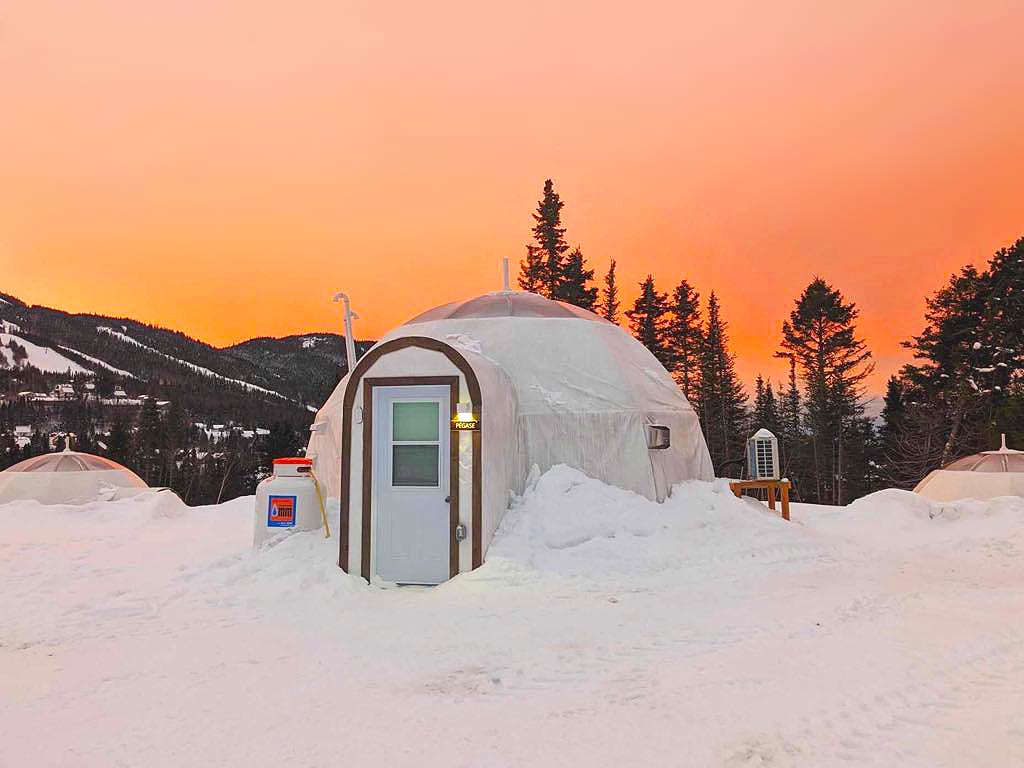 The exterior of a geodesic dome is set against a vibrant orange sunset, surrounded by a snowy landscape and evergreen trees. Snow covers the ground, and additional domes can be seen in the background, highlighting the tranquil winter setting.