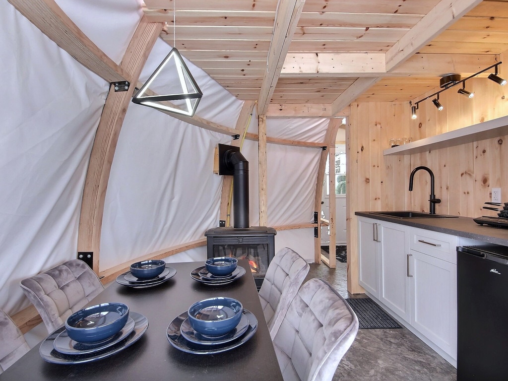 A dining area features a dark table set with navy blue plates and plush gray chairs. A modern triangular light fixture hangs from the ceiling. A propane fireplace is visible, providing warmth, while white walls and natural wood accents create a cozy atmosphere.