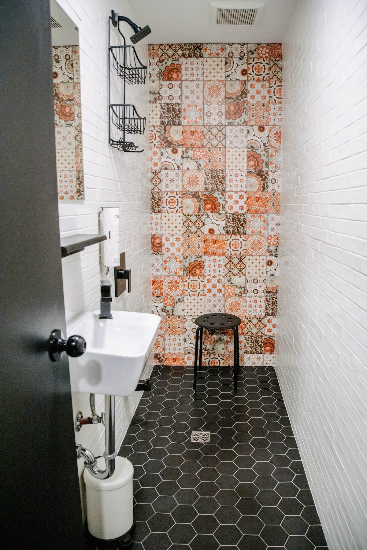 A bathroom is displayed featuring a modern sink with sleek fixtures. A decorative wall with patterned tiles adds visual interest. A small black stool is positioned on the hexagonal tiled floor, and wall-mounted shelving holds toiletries. The overall space is well-lit and designed for efficient use.