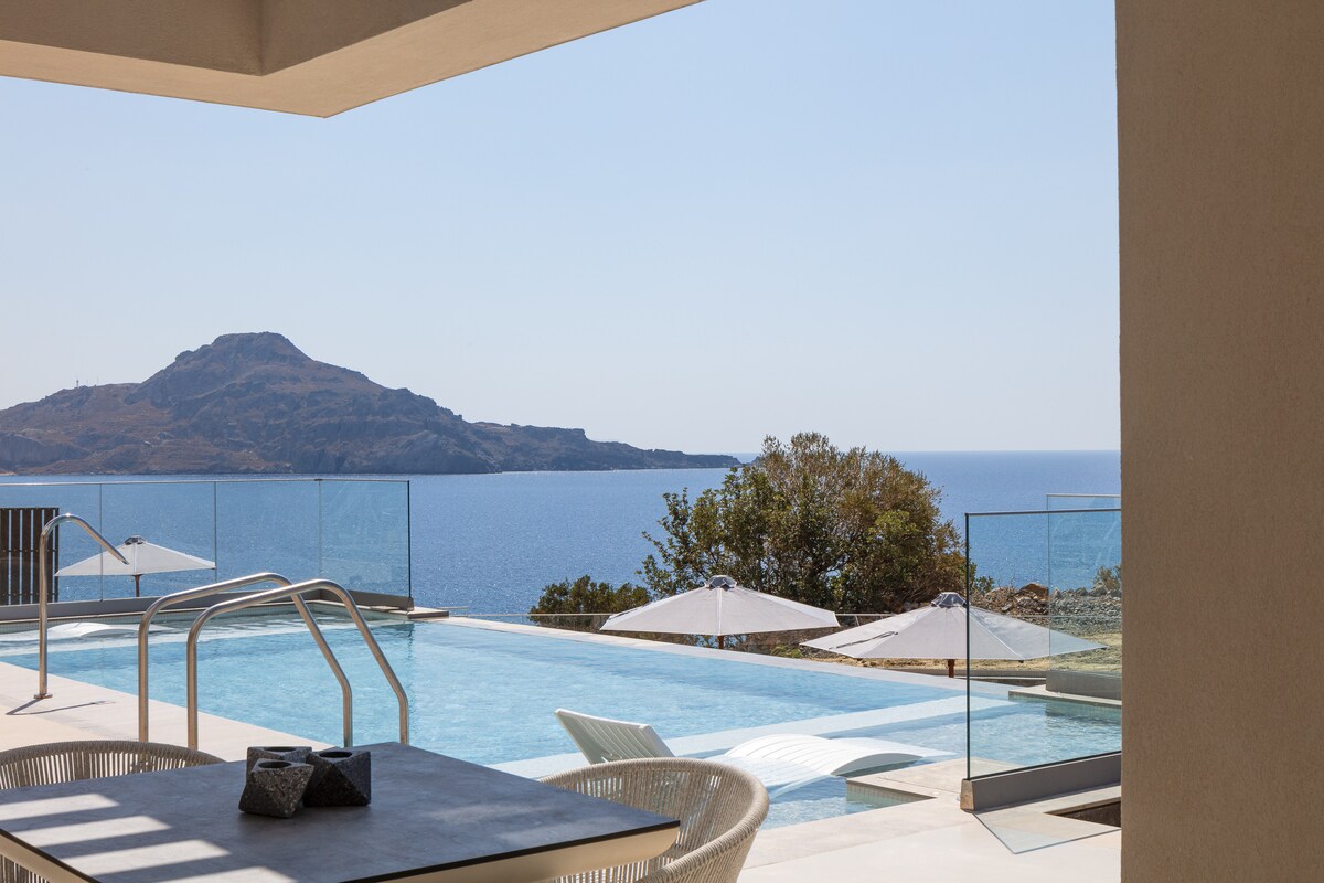A spacious outdoor area features a private swimming pool with clear blue water, accompanied by lounge chairs. Transparent glass panels provide unobstructed views of the sea and distant hills, while sun umbrellas offer shade. The scene is set in a bright, sunny environment.