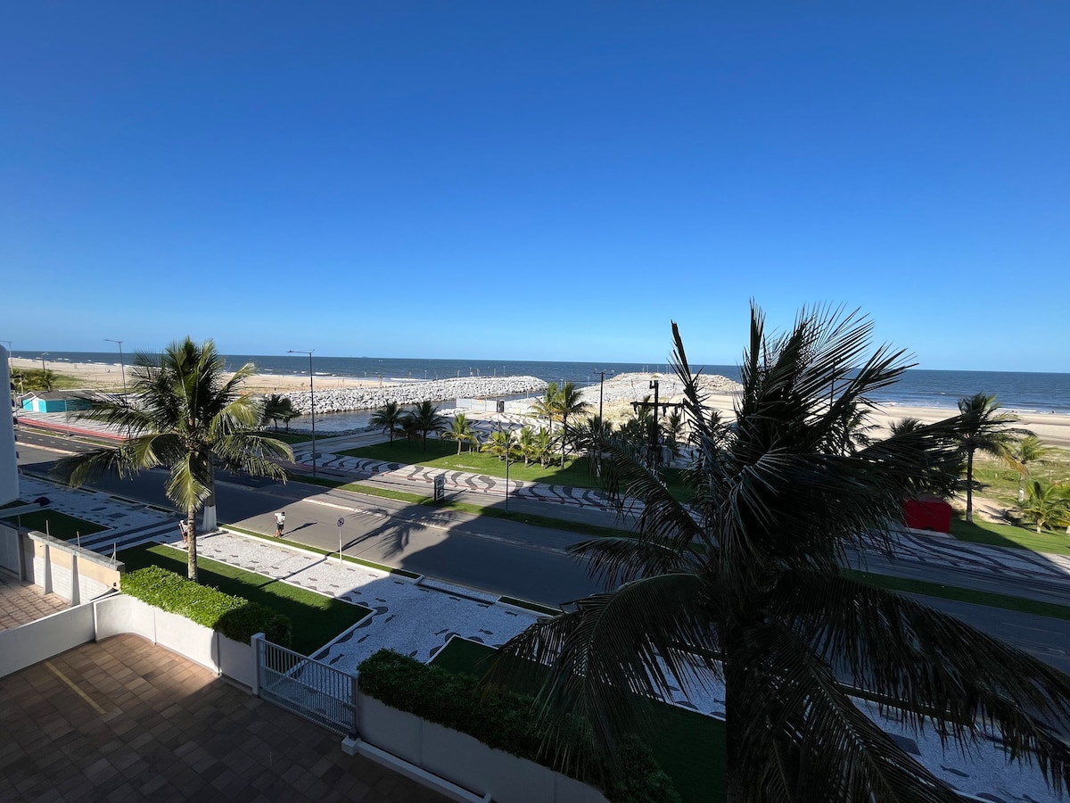 A scenic view of the beach is captured from a balcony, showcasing the sandy shoreline and the ocean. Palm trees are visible in the foreground, framing the image, while the clear blue sky highlights the open landscape.