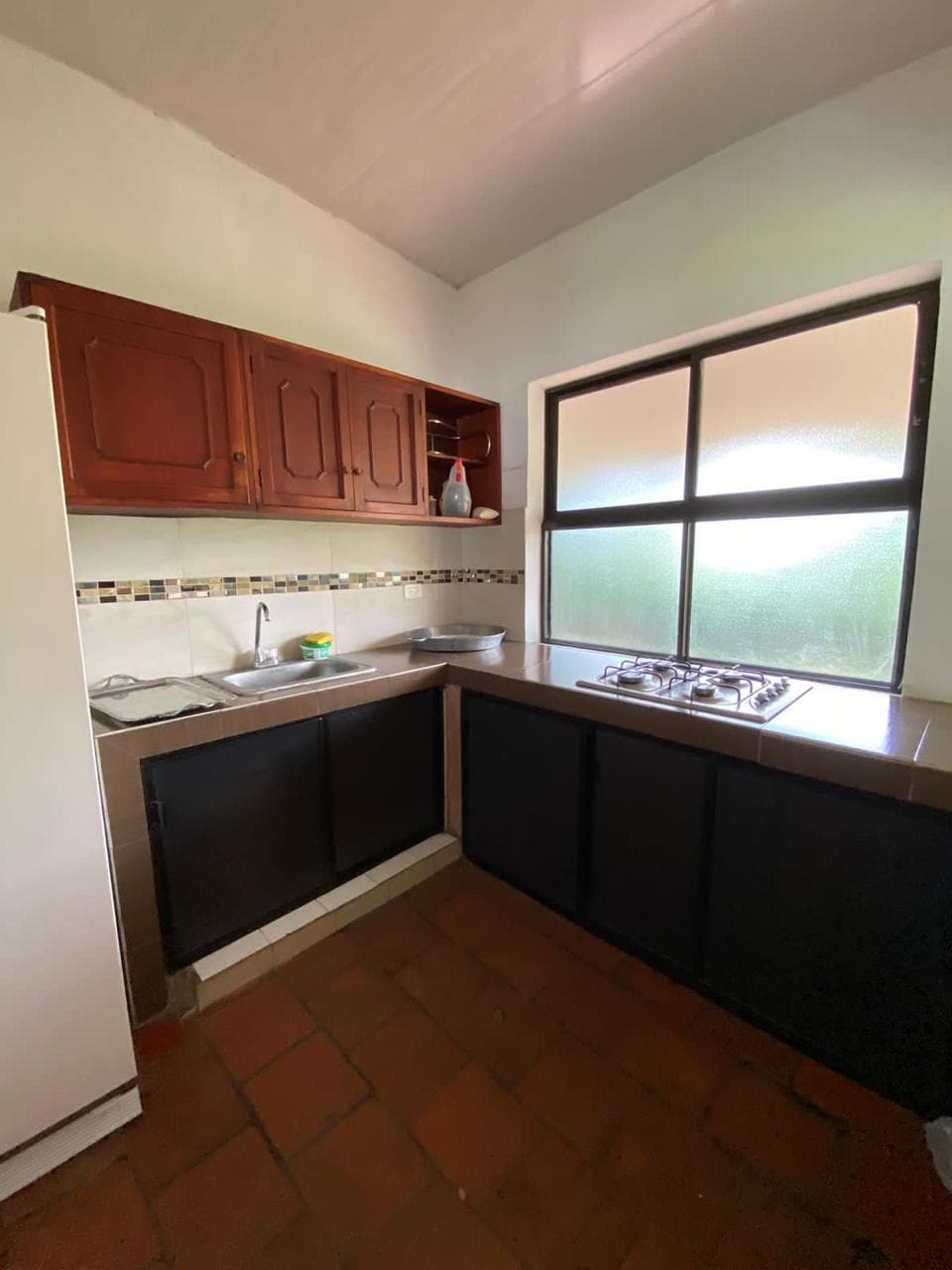 A compact kitchen area is displayed with wooden cabinets providing storage above the countertop. A stainless steel sink and a gas stove are featured alongside a window that allows natural light to enter, illuminating the space.