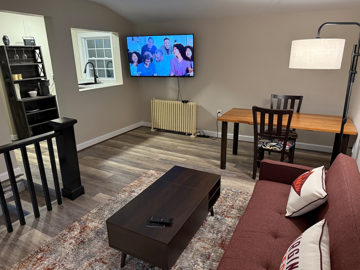 A comfortable living area features a red couch facing a wall-mounted television. A dining table with chairs occupies one corner, next to a radiator and a window. The warm-toned flooring is complemented by an area rug, creating a cozy atmosphere.