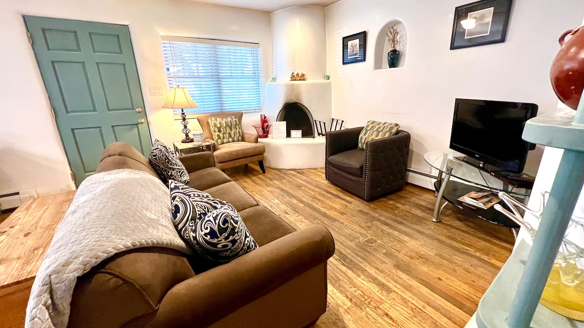 A comfortable living area is showcased with a plush brown sofa and accent chairs. A kiva fireplace is set against the wall, complemented by a small television on a glass stand. Natural light filters through a window adorned with blinds.