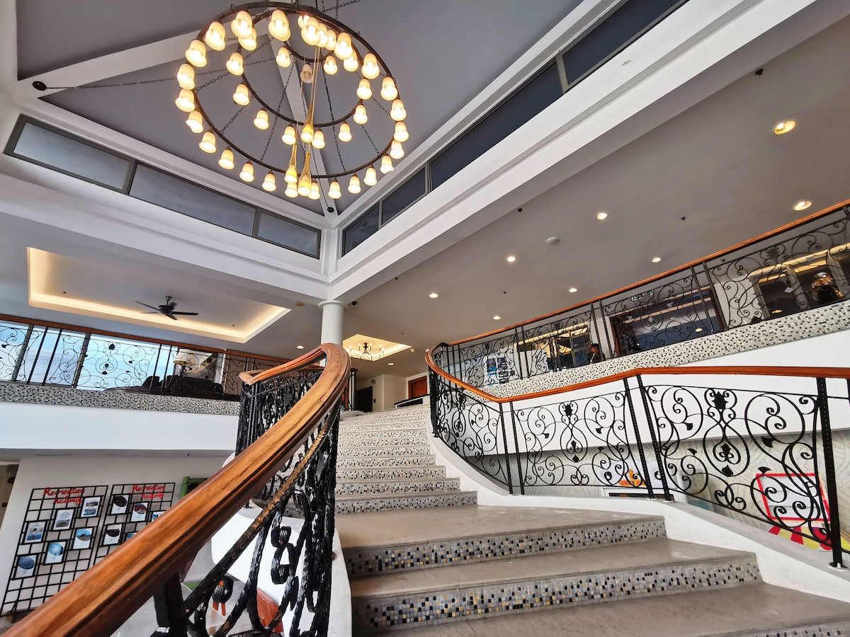 A grand staircase features an ornate railing and a circular chandelier overhead, illuminating the spacious lobby area. Natural light filters through large windows, enhancing the airy atmosphere. Decorative wall art and comfortable seating can be seen in the background.
