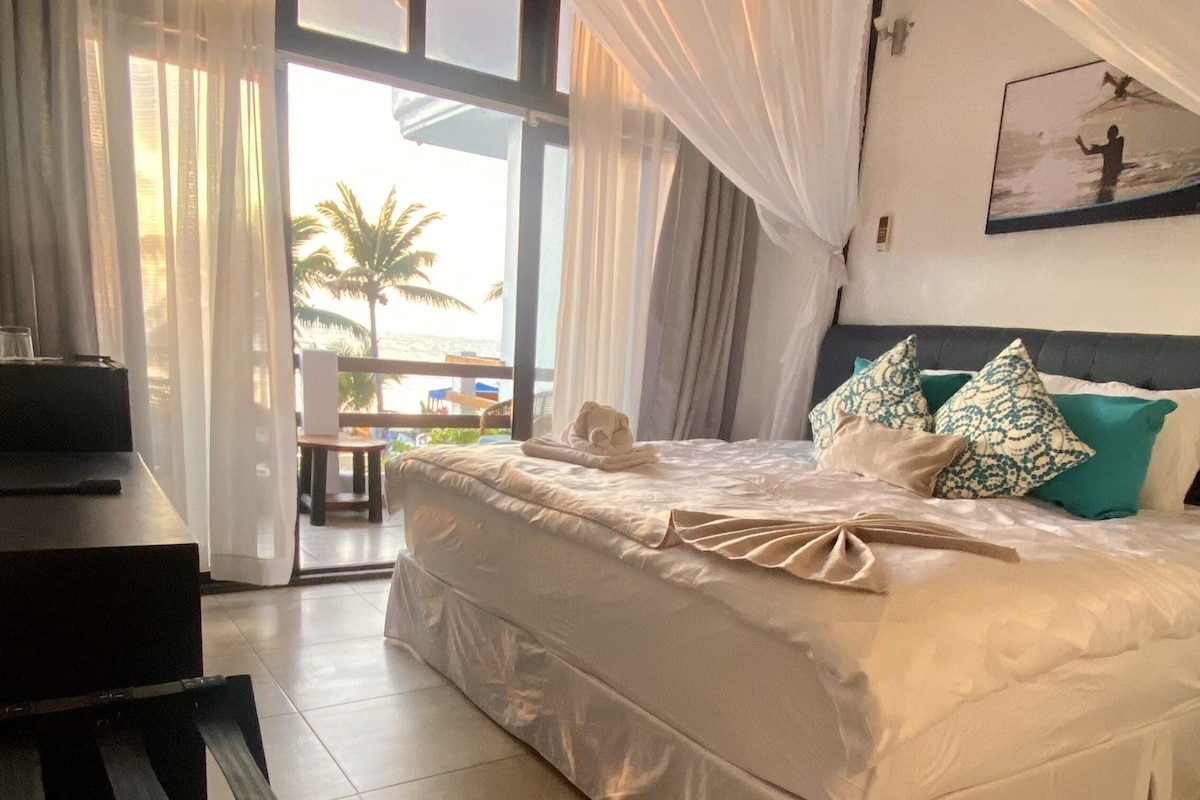 A spacious bed is adorned with decorative pillows and a folded towel, positioned beside large glass doors. Sheer curtains gently frame the entryway, allowing natural light to fill the room, while palm trees and outdoor seating are visible beyond the balcony.