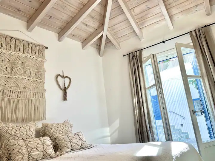 A bright bedroom features high wooden ceilings and a textured wall hanging. Natural light filters through two large windows with elegant curtains, illuminating a spacious bed adorned with decorative pillows. A cozy and inviting atmosphere is created, offering a connection to the outdoor space.