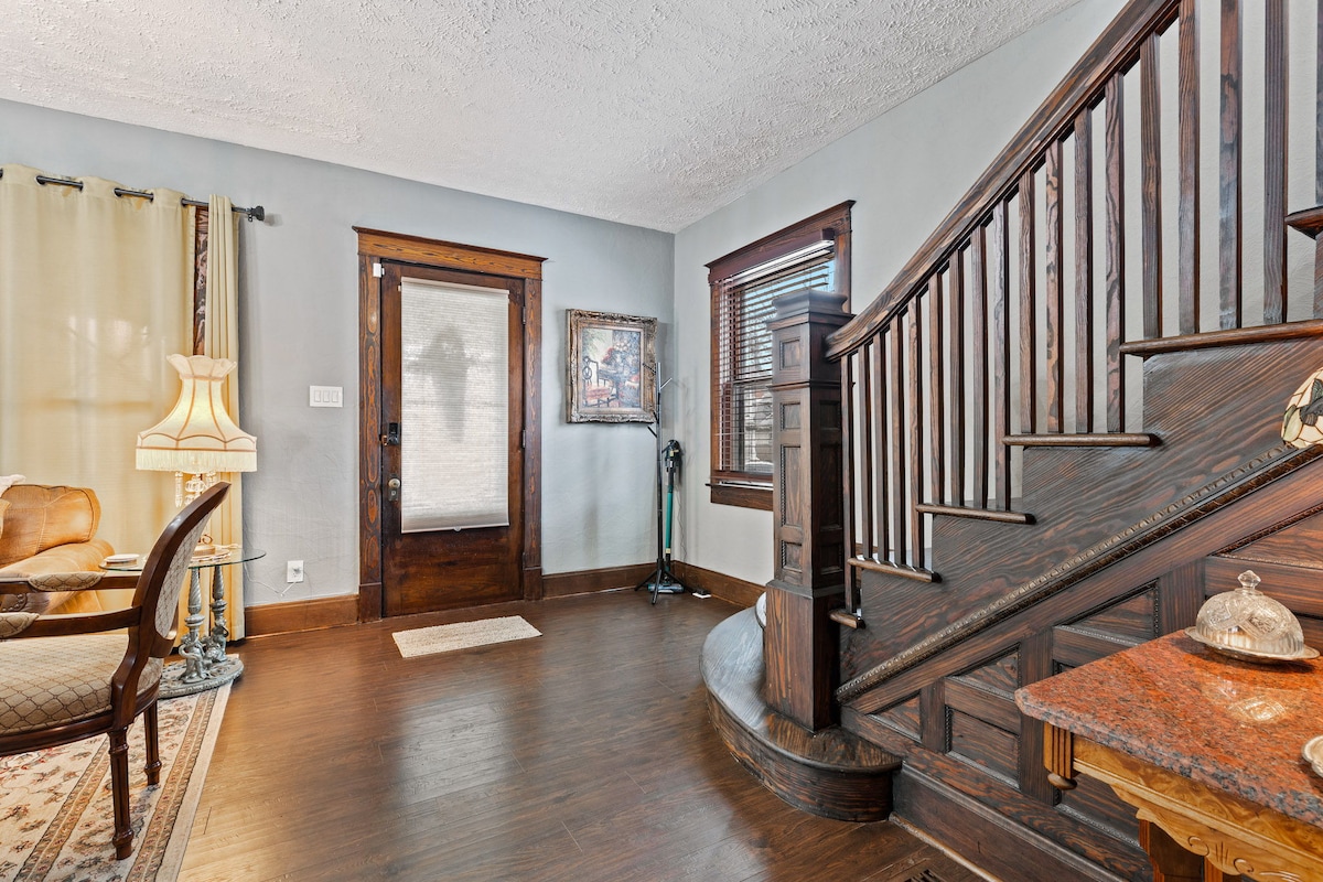 An elegant entryway is showcased, featuring rich wooden accents and a sweeping staircase. Natural light filters through the front door and curtains, illuminating the inviting space. A comfortable seating area is presented, complemented by tasteful decor elements and a patterned area rug.