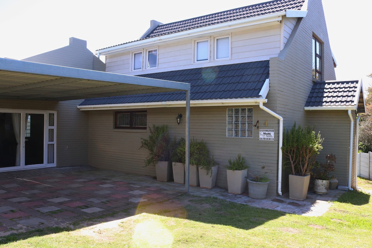 The exterior of the holiday home displays a modern two-story design featuring light grey siding and a pitched roof. A covered patio area is visible, adorned with potted plants, while a spacious lawn extends in front, providing a welcoming entryway.