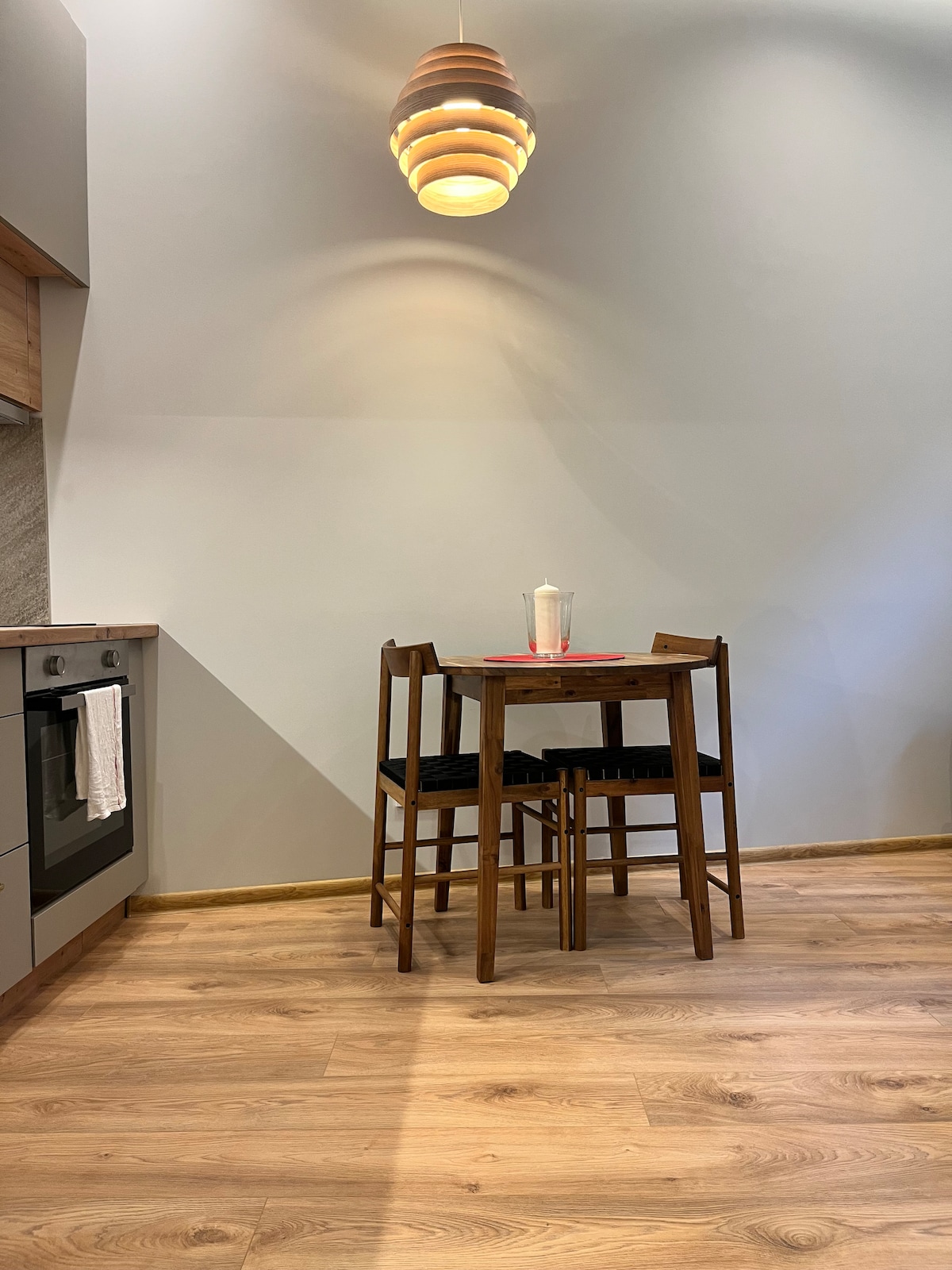 A cozy dining area is highlighted by a wooden table surrounded by four high-backed stools. A sleek pendant light hangs above, casting a warm glow. The kitchen is equipped with modern appliances, and light wood flooring contributes to a bright and inviting atmosphere.