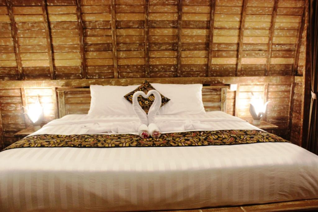 A spacious bed is adorned with a decorative blanket and accented by heart-shaped towel art. The wooden walls provide a rustic ambiance, while soft lighting creates a serene atmosphere. Two bedside lamps add to the cozy setting.
