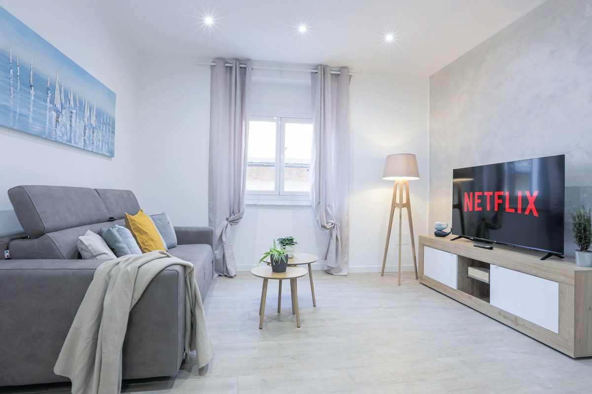 A bright living room featuring a comfortable gray sofa with accent pillows and a soft throw. A light wood TV stand displays a smart TV with Netflix. Natural light enters through the window, complemented by light curtains and overhead recessed lighting.