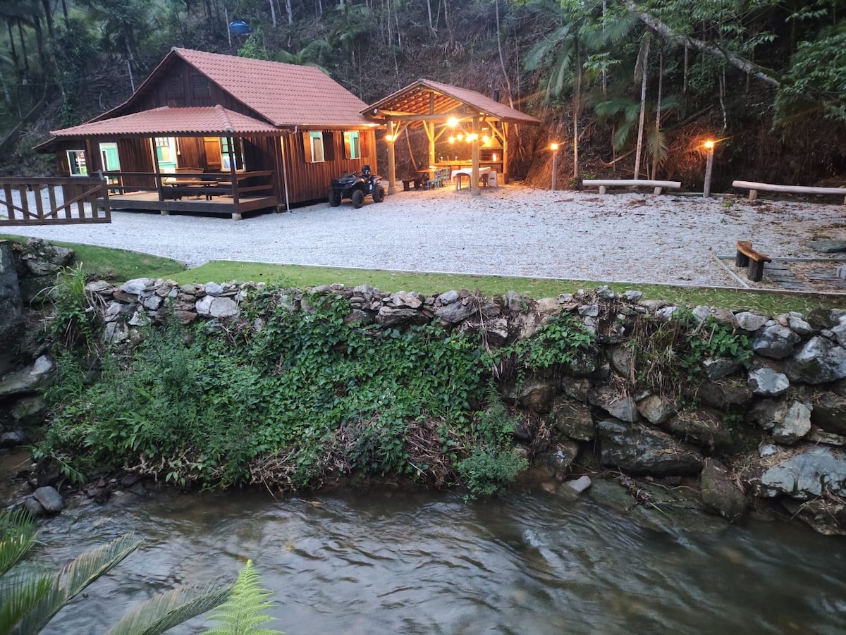 A charming cabin is situated alongside a gently flowing stream, surrounded by lush greenery. Outdoor seating areas are visible, with soft lighting illuminating a covered space. The gravel driveway leads to the cabin, enhancing the peaceful natural setting.