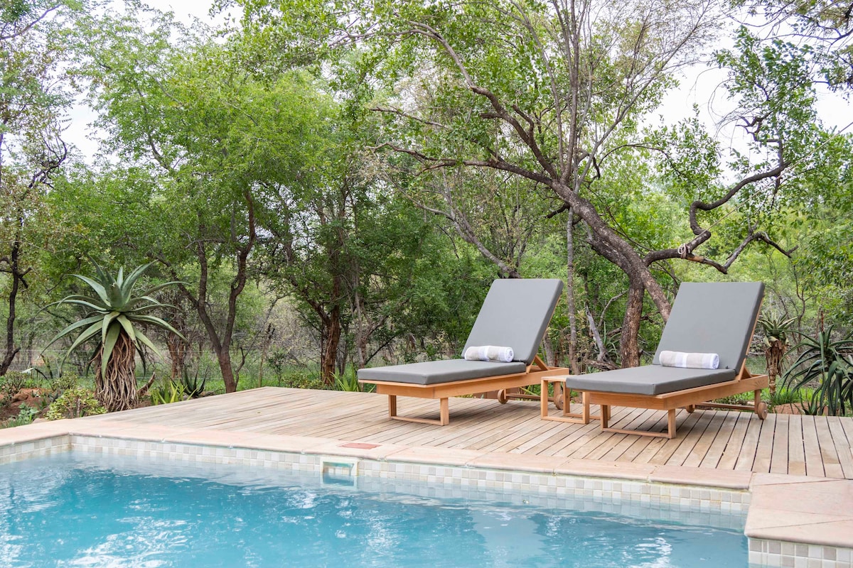 Two lounge chairs with gray cushions and rolled towels are positioned beside a swimming pool. A wooden deck surrounds the pool, offering a natural view of lush greenery and shrubs, enhancing the outdoor relaxation area.