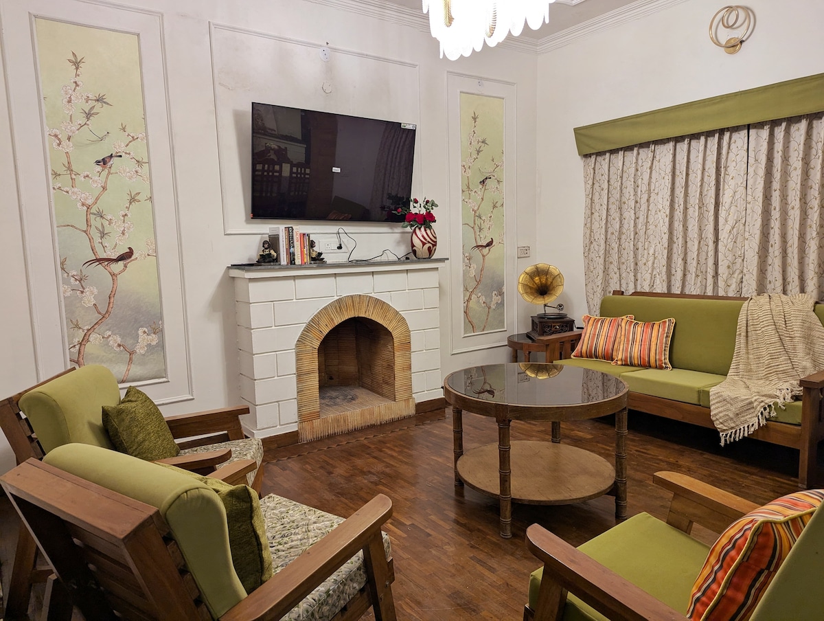 A cozy living area features two green sofas and a circular wooden table at the center. A white brick fireplace adds warmth, while a wall-mounted TV is visible above. Two decorative wall panels showcase floral designs, and a window is adorned with soft curtains.