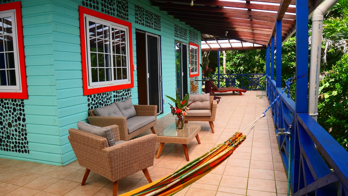 A spacious terrace features comfortable seating arrangements with a glass table and decorative plants. Brightly colored walls complement the natural surroundings. A hammock is positioned nearby, inviting relaxation, while vibrant outdoor furniture enhances the welcoming atmosphere.