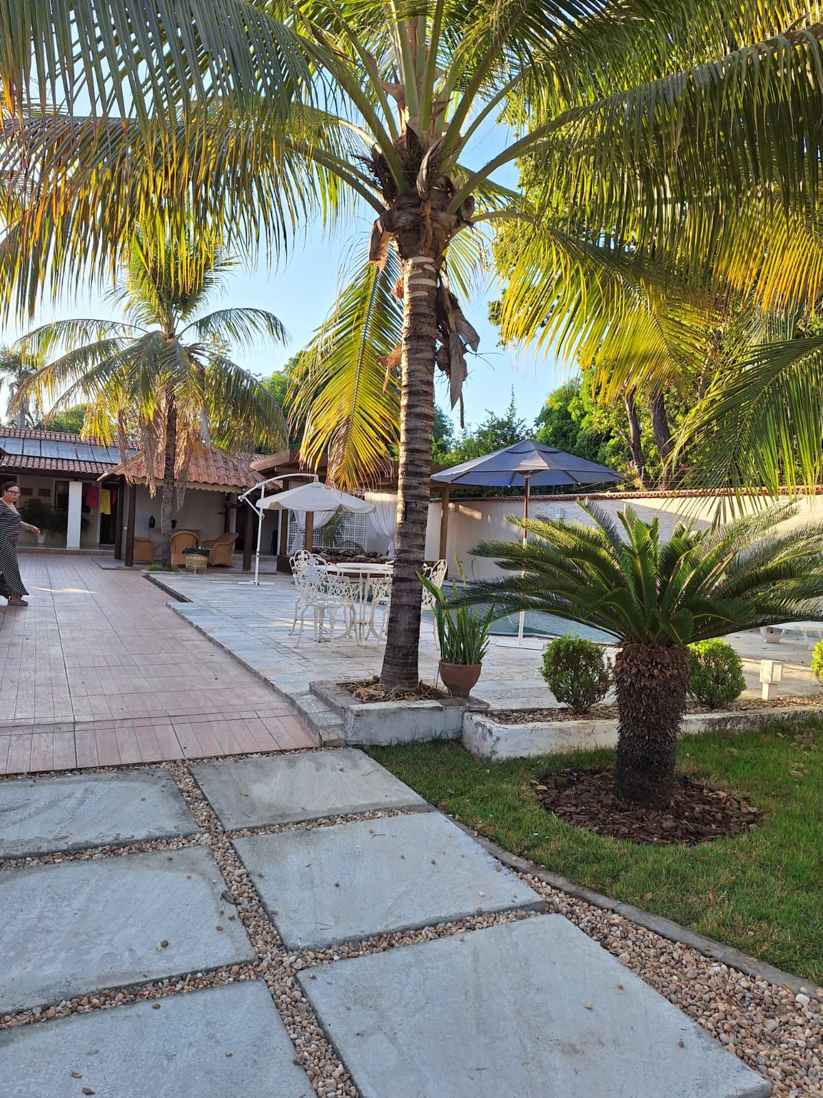 The outdoor area features a well-kept courtyard with a mix of paved and grassy sections. Palm trees provide shade, while a seating area with a table and chairs is visible. A gazebo can be seen in the background, enhancing the space's inviting atmosphere.