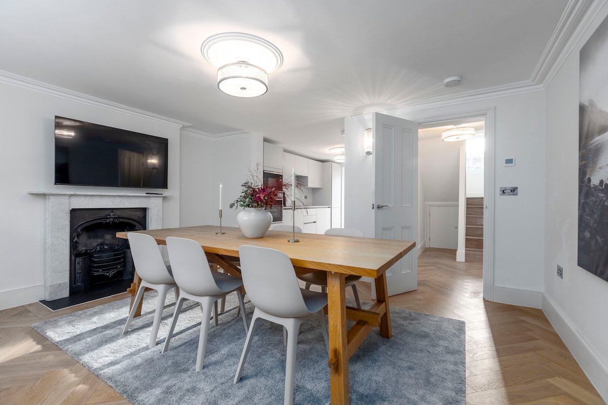 A dining area features a wooden table surrounded by six chairs, set on a soft rug. A mounted flat-screen television is above a marble fireplace, while a doorway leads towards the kitchen. Warm lighting from the ceiling fixtures enhances the bright, neutral-toned decor.