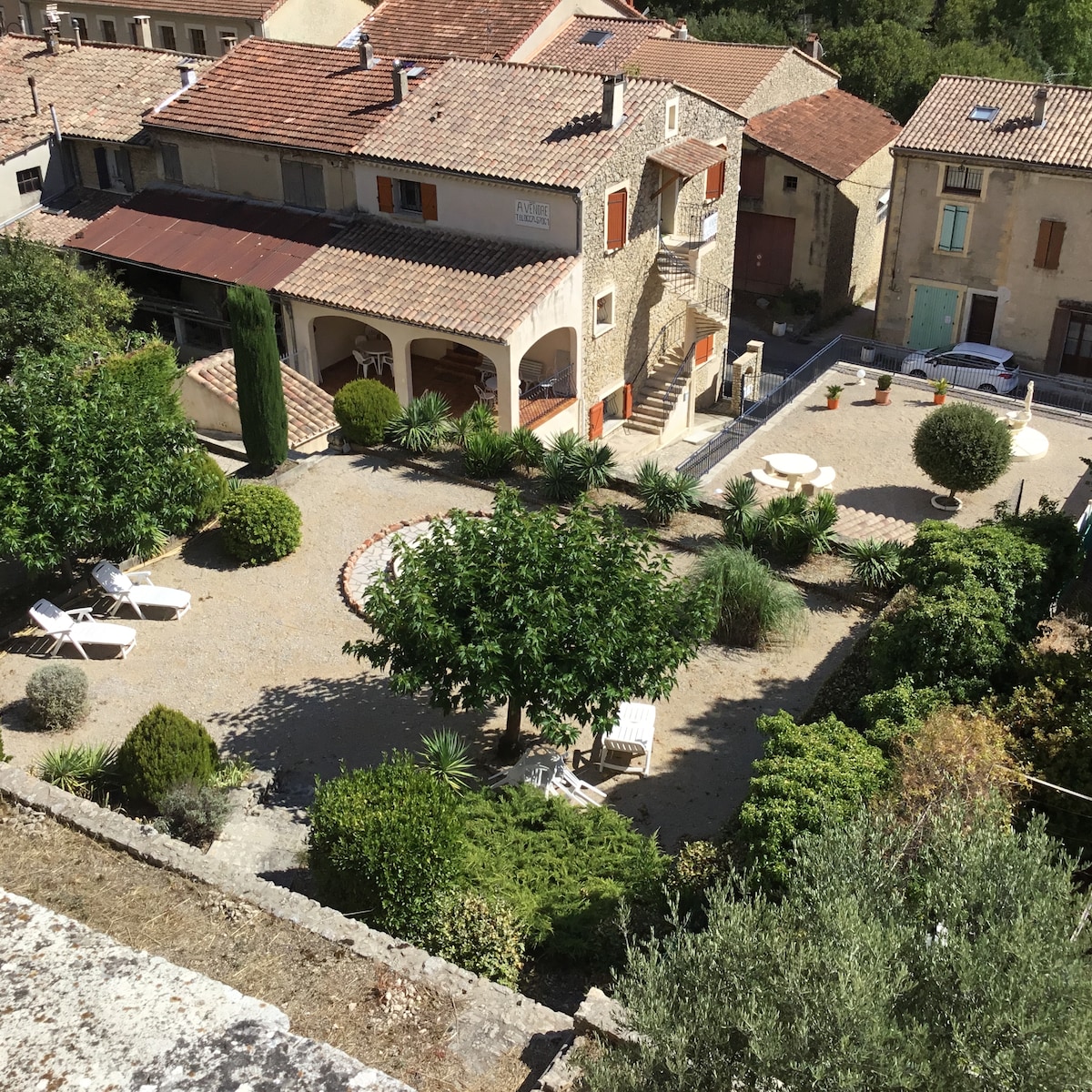 An expansive garden is depicted, featuring a variety of plants and trees. Lounge chairs are arranged near a circular path, with tables placed among the greenery. The surrounding buildings reflect a rustic architectural style, providing a tranquil setting within the landscape.