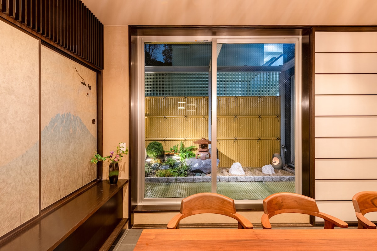 A serene indoor space is highlighted by a large glass window revealing a neatly arranged garden. A traditional stone lantern and lush greenery can be seen outside, enhancing the tranquil atmosphere. Wooden dining chairs are positioned around a robust wooden table.