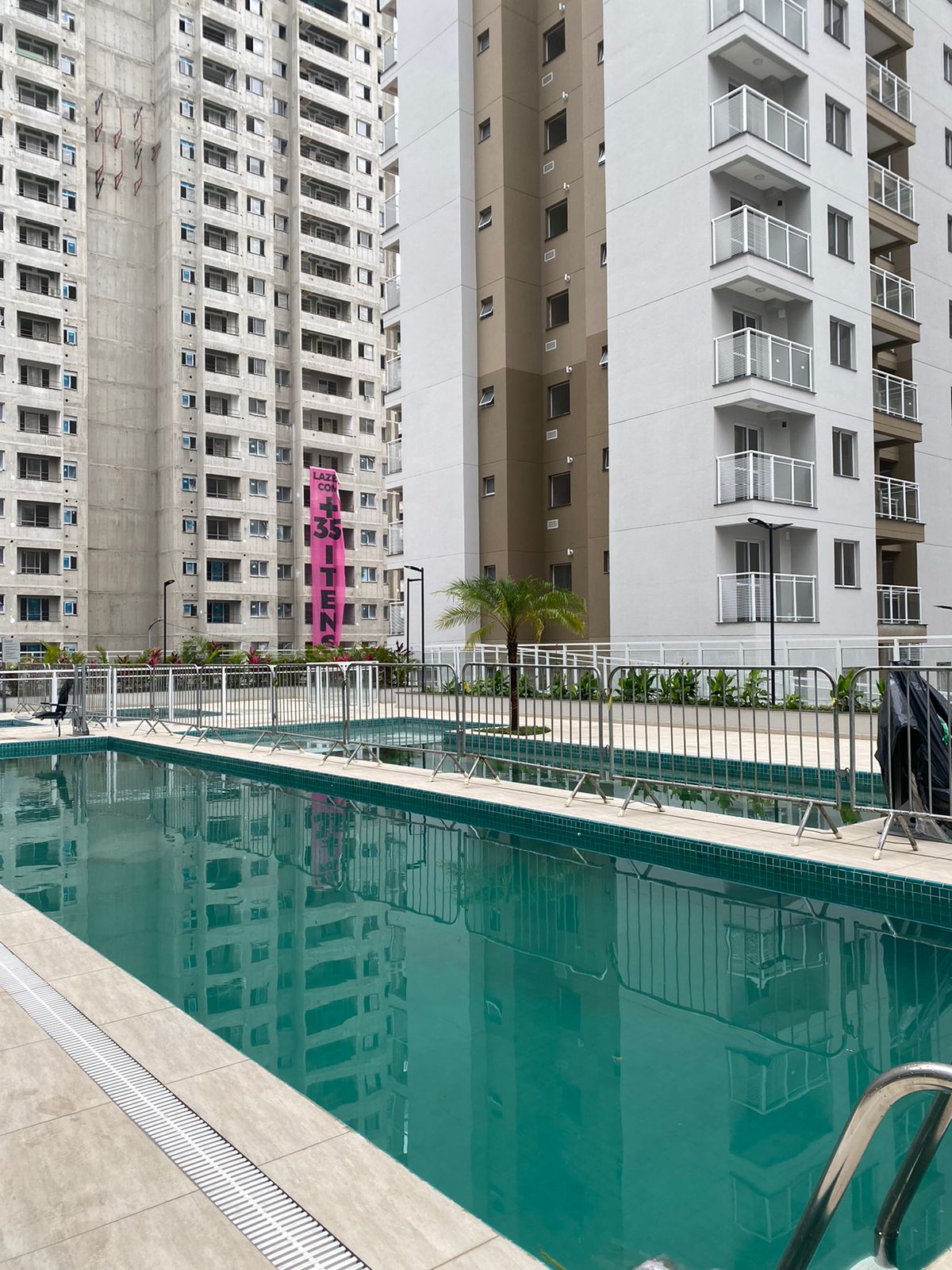 The image displays a swimming pool surrounded by modern buildings, featuring a clear blue water surface. The pool area includes lounge space, with fencing and landscaping visible. Surrounding structures are seen in the background, showcasing a mix of colors and architectural styles.
