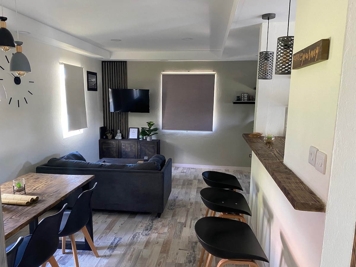 An open-concept living and dining area features a dark sofa facing a wooden dining table. Bar stools line a counter, with light fixtures hanging overhead. Two windows are dressed with roller shades, allowing natural light to filter in, while a small plant is visible on the counter.