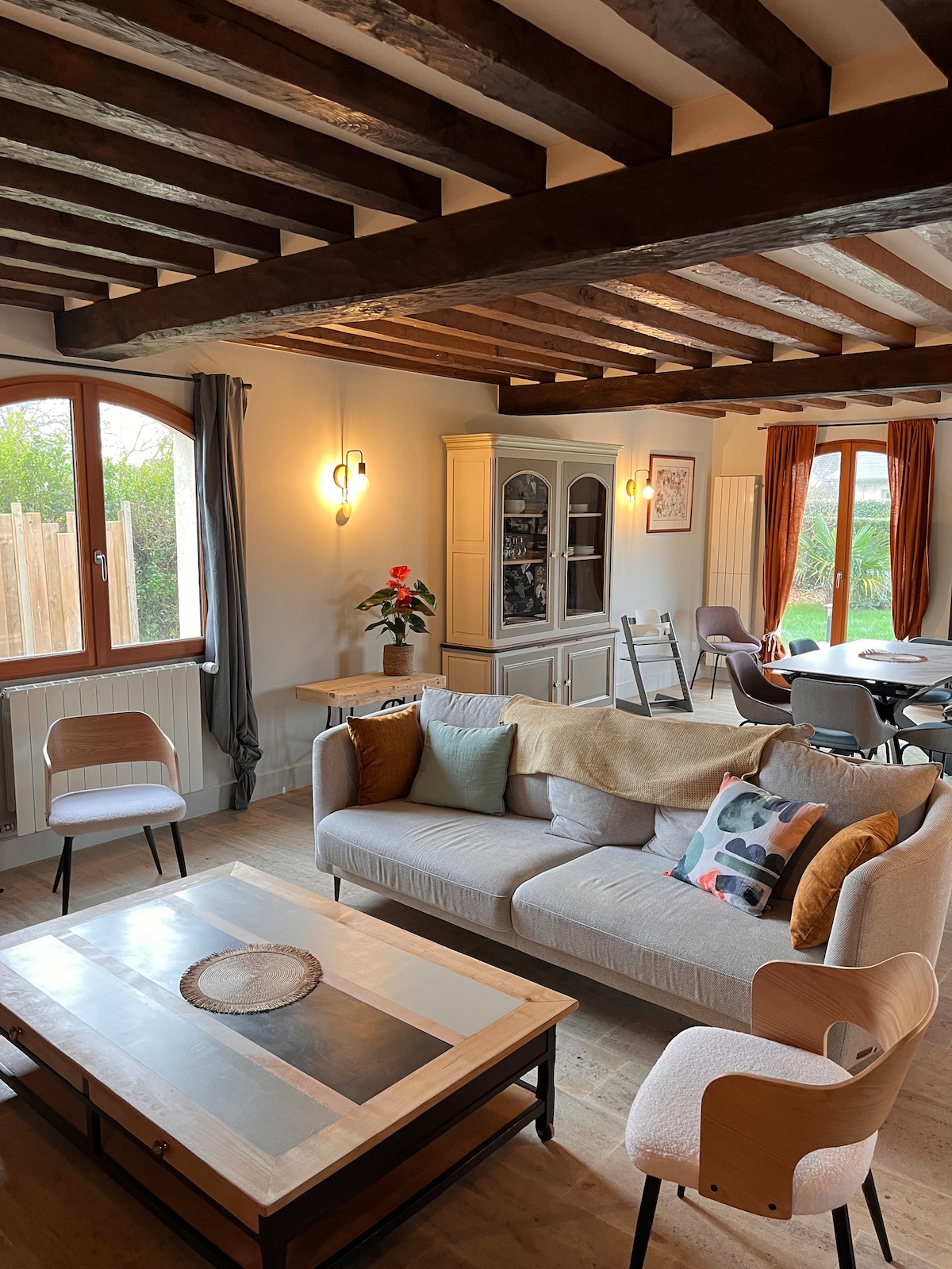 The spacious living area features wooden beams overhead and natural light streaming through large windows. A comfortable sofa is adorned with colorful cushions, while a coffee table rests in the center. A dining area with modern chairs and a sideboard complements the warm ambiance of the space.