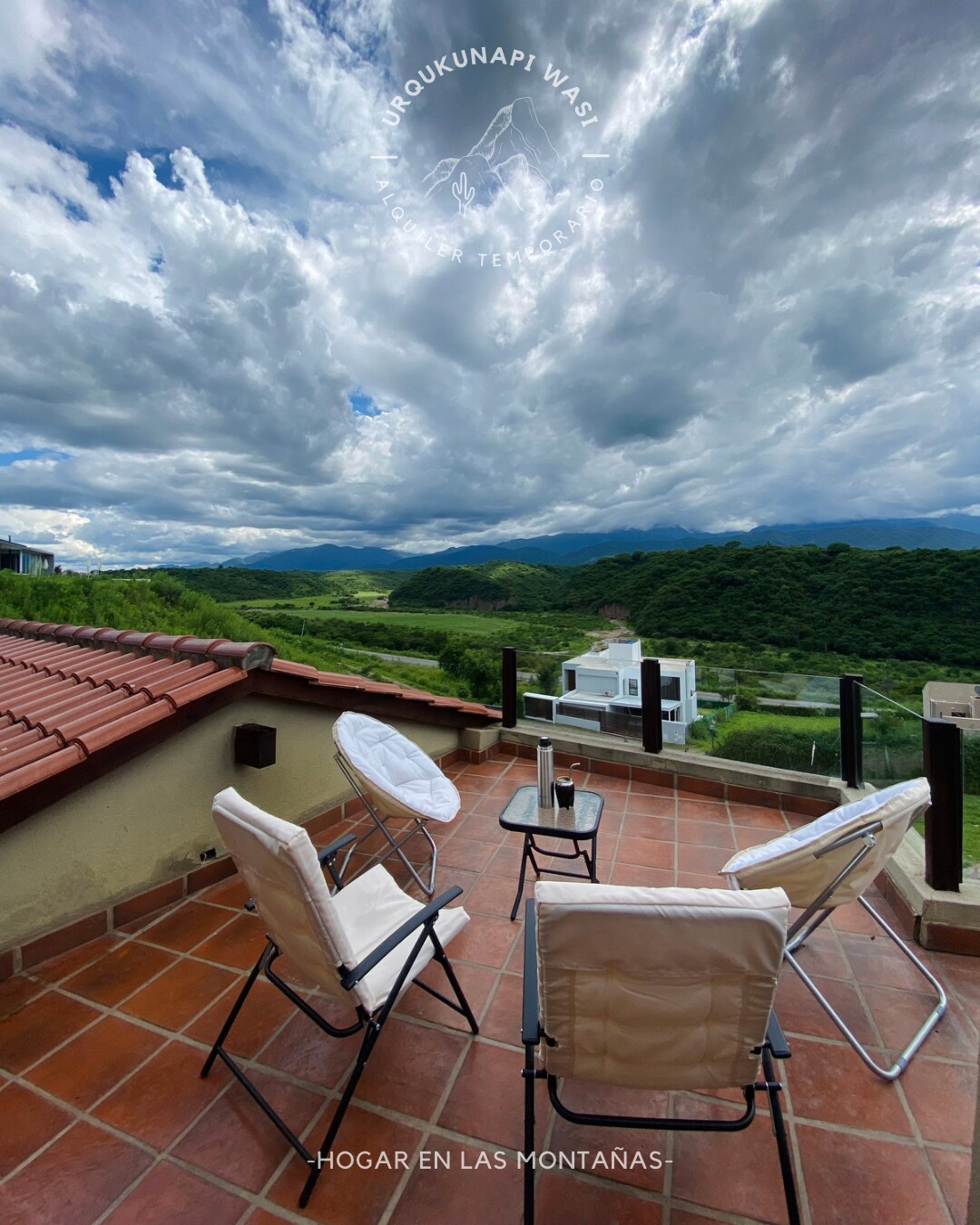A rooftop terrace is showcased with terracotta tiles and glass railing, providing views of the surrounding mountains and valleys. Two white lounge chairs and a small table are positioned for relaxation, inviting guests to enjoy the scenic landscape under a dramatic sky filled with clouds.