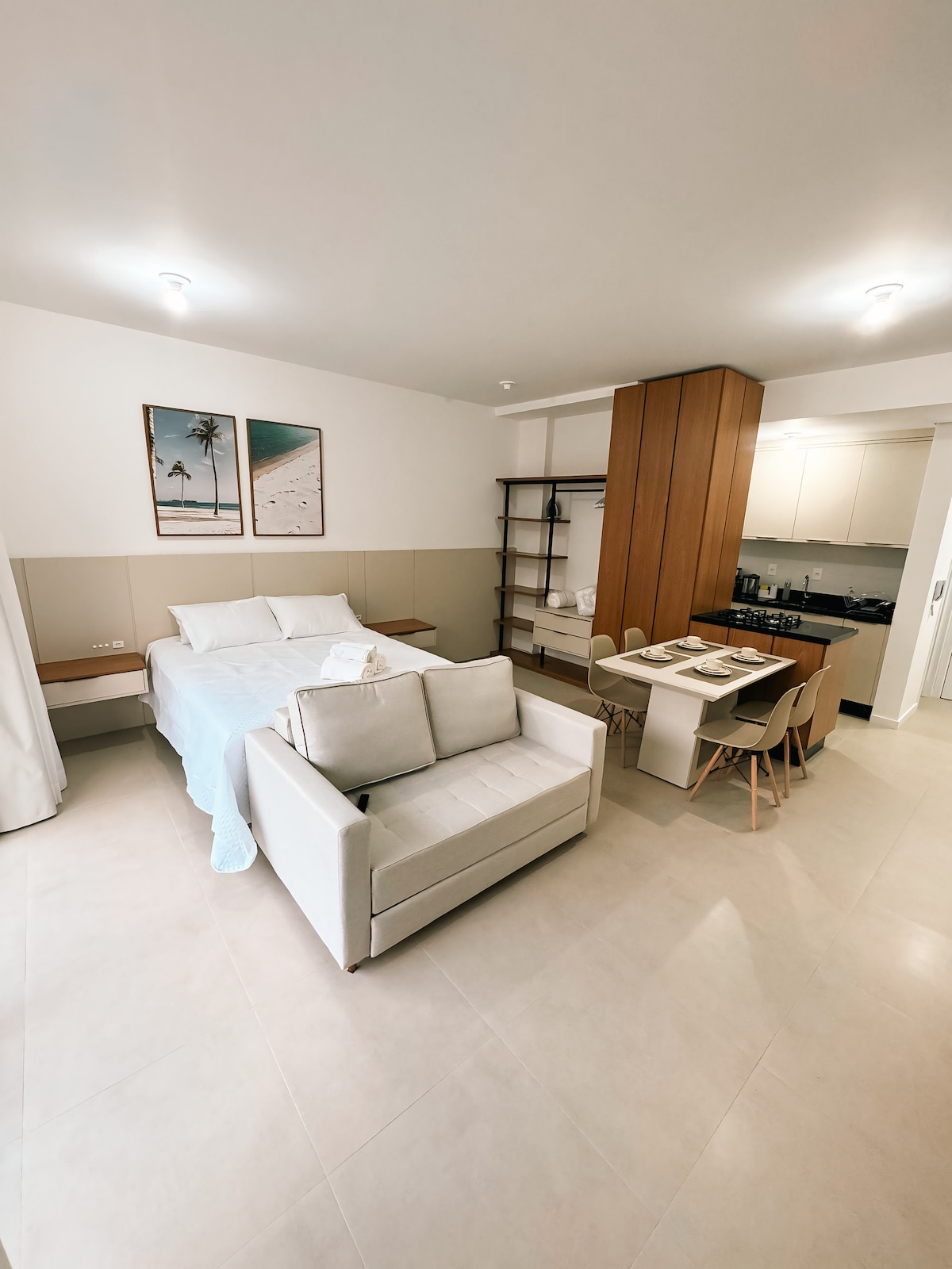 A modern studio apartment is displayed, featuring a spacious bed with white linens and a soft sofa facing a small dining area. Natural light illuminates the light-colored tile floor, while a simple wooden shelf and a kitchenette are visible in the background.