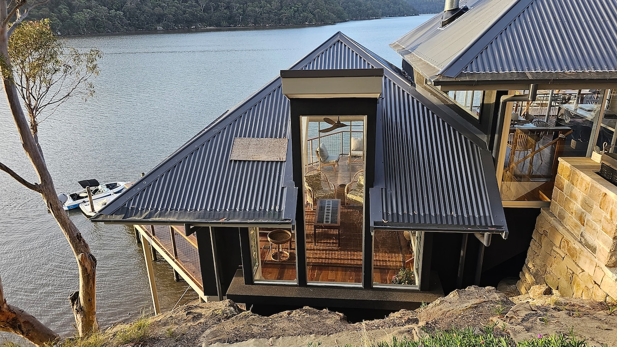 A unique water access property is visible with a sloped roof and expansive glass windows, offering views of the surrounding river. The deck extends towards the water, and a boat is docked nearby, providing easy access for outdoor activities and relaxation.