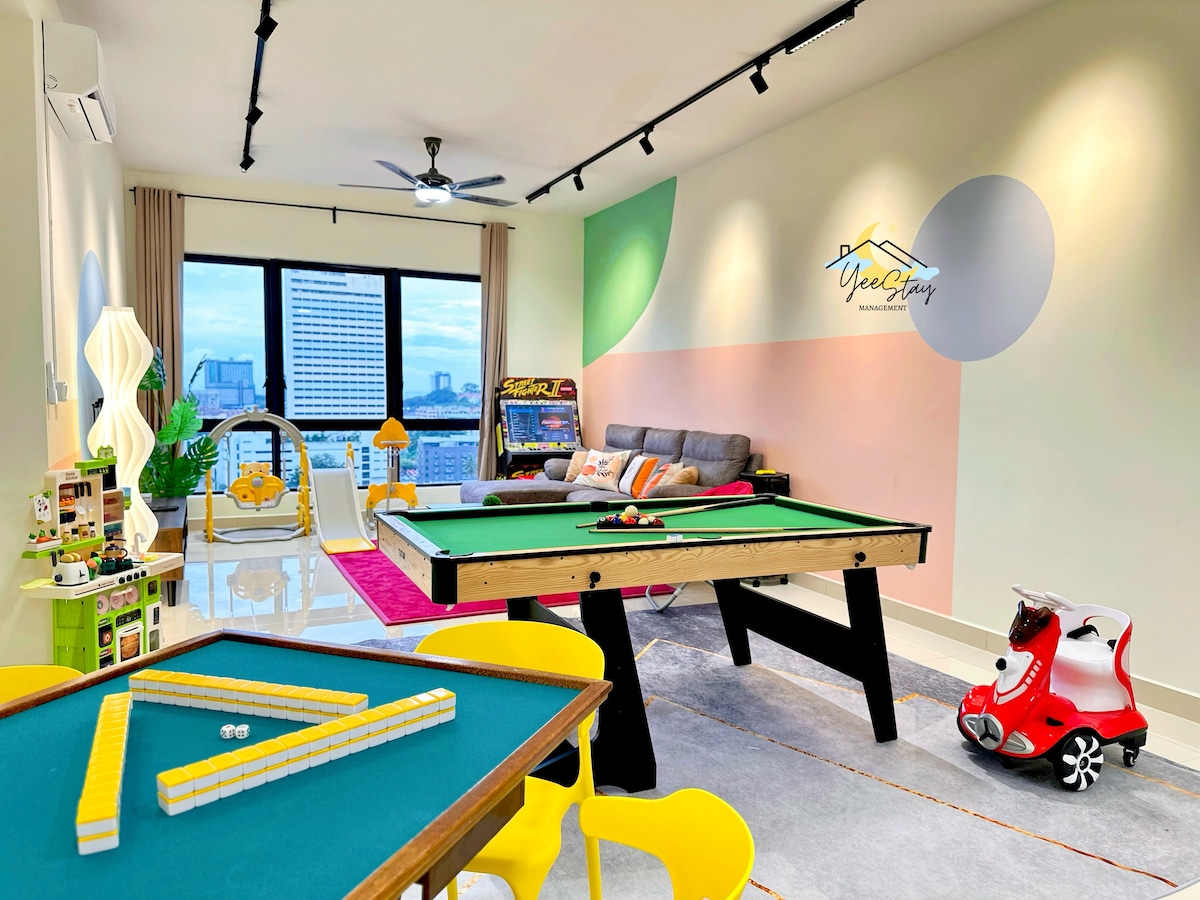 A vibrant entertainment space features a green pool table and a colorful gaming console cabinet. A cozy seating area is positioned near a large window, allowing natural light to illuminate the playful decor. Yellow chairs and a whimsical toy vehicle add family-friendly appeal.
