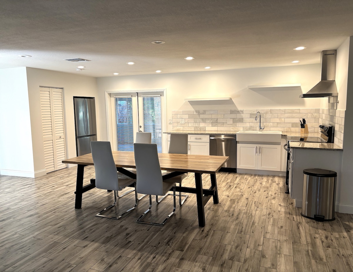 An open-concept kitchen and dining area features a large wooden dining table surrounded by four sleek chairs. A modern kitchen with stainless steel appliances, white cabinetry, and a tiled backsplash is visible. Ample natural light streams in through the glass doors leading to the outside.