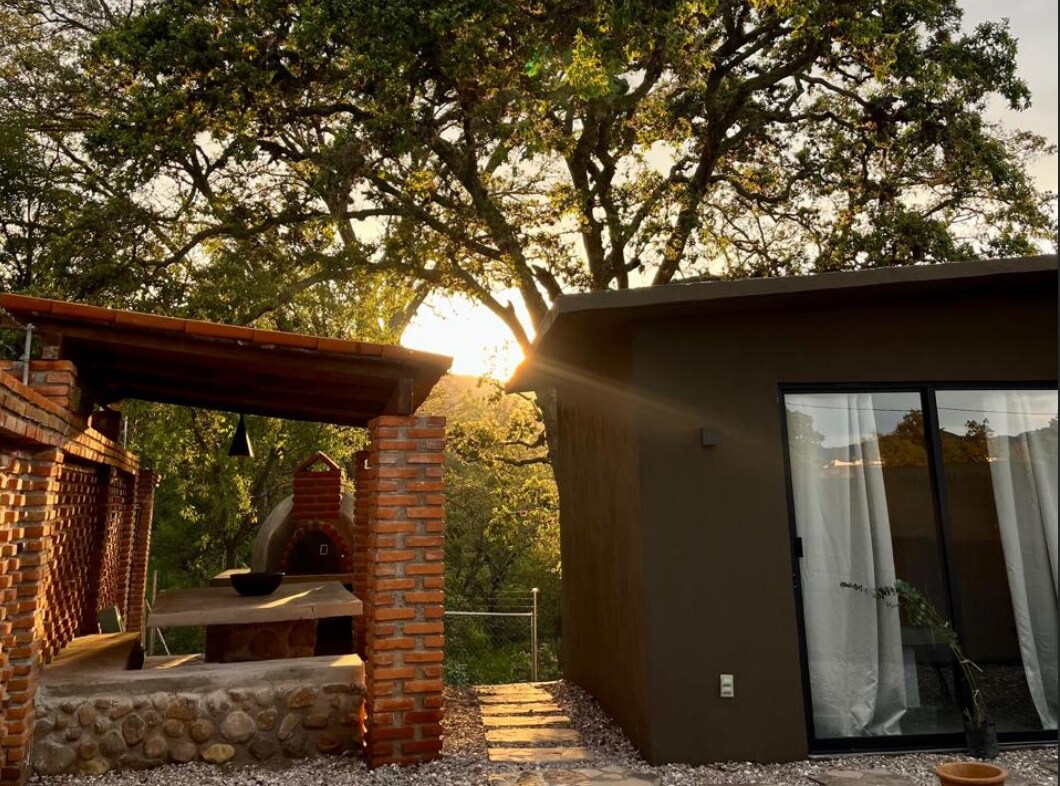 A charming outdoor kitchen area features a traditional oven constructed from brick, with a stone surface for food preparation. A modern dwelling stands nearby, framed by the warm glow of sunset filtering through the leaves of a large tree. A peaceful ambiance is created.