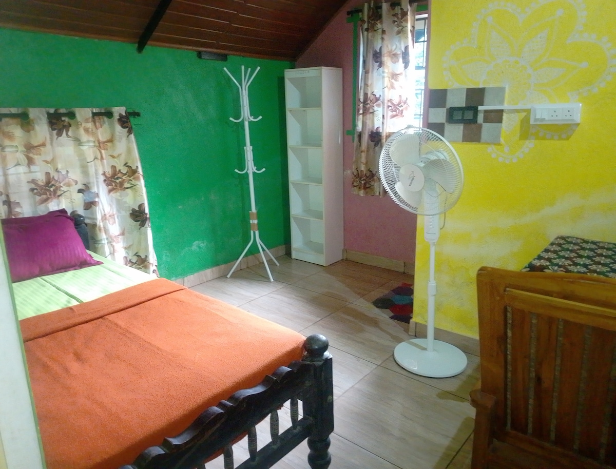 A vibrant room features a single bed with an orange blanket and accent pillows. A small table and chairs are present, alongside a white fan and a coat rack. Colorful walls in green and yellow add character, while a window is adorned with floral curtains.