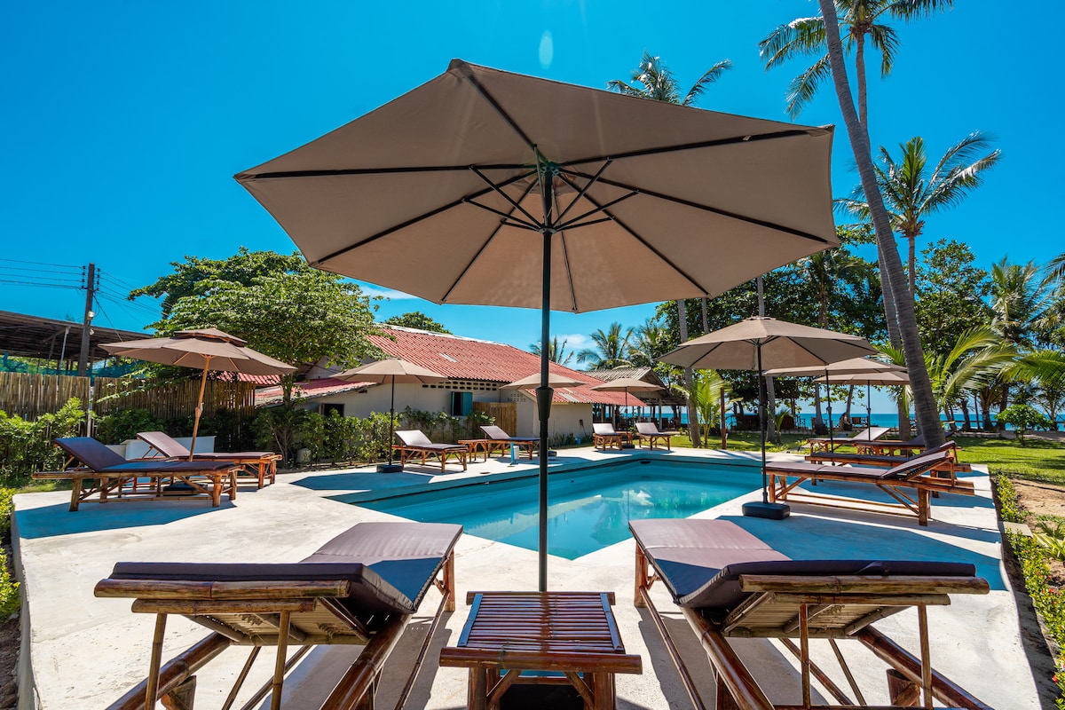 A serene outdoor area features a swimming pool surrounded by lounge chairs and large umbrellas. Lush greenery and palm trees create a refreshing atmosphere, while clear blue skies enhance the bright, inviting space. A spacious deck offers room for relaxation and enjoyment.