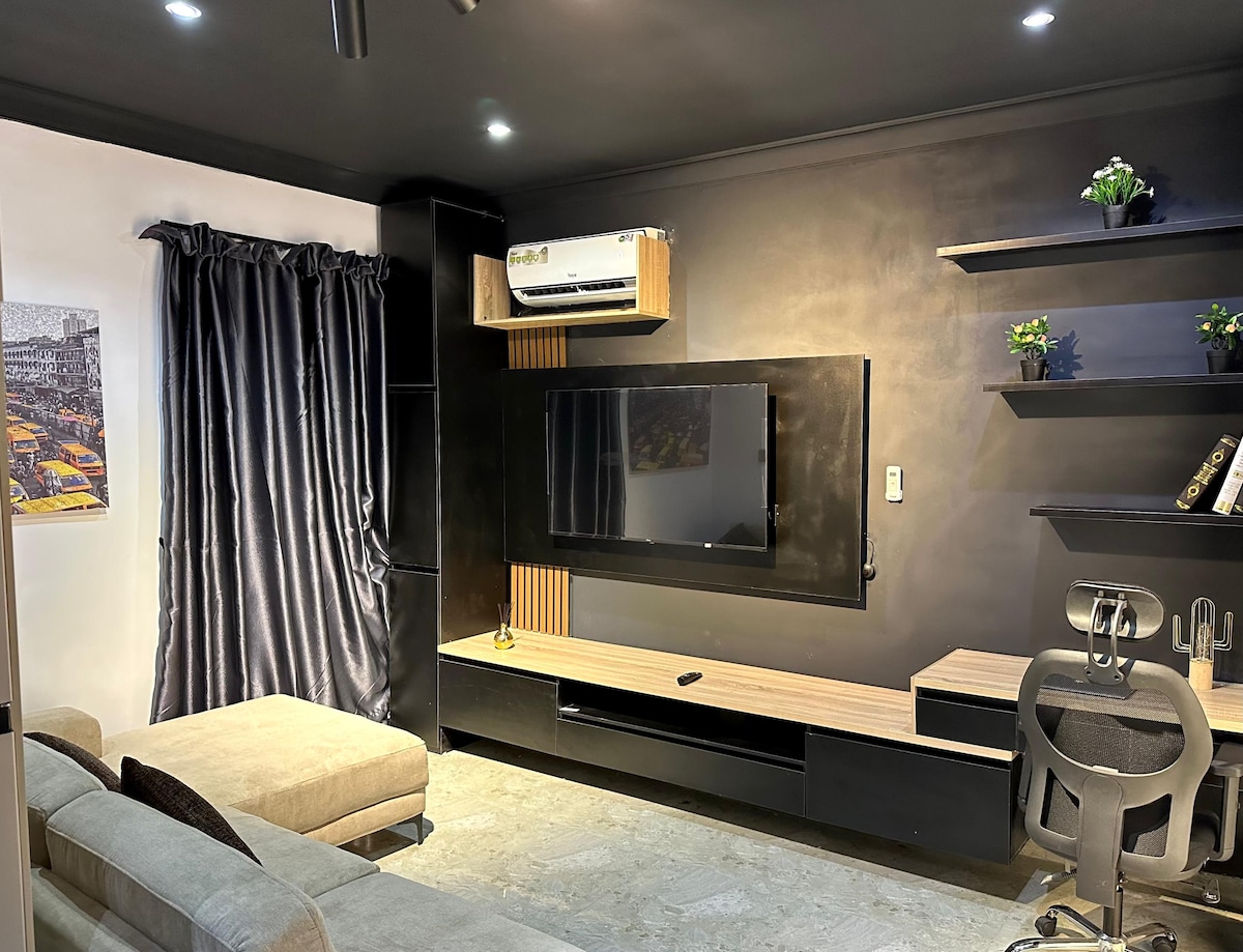 A modern living space features a comfortable sofa and a wall-mounted television. Dark curtains frame the window, while sleek shelves showcase decorative items. A small desk with a chair is positioned nearby, and a wall-mounted air conditioning unit is visible, providing convenience.