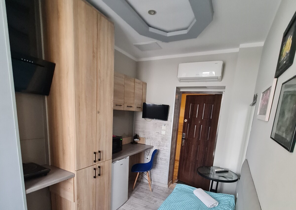 A compact studio room features a spacious bed and a wooden closet. A small desk with a blue chair is positioned alongside a wall-mounted television. Natural light illuminates the space, accentuated by an air conditioning unit and framed artwork. The entrance door is visible in the background.