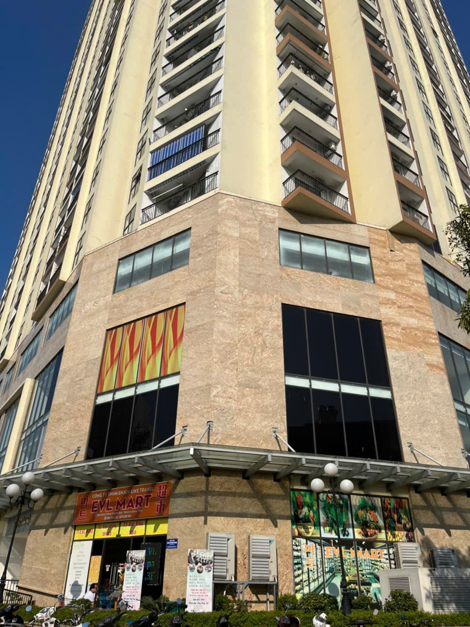 A modern high-rise building features a combination of glass and stone facades, with large windows encouraging natural light. The ground level includes a well-lit entrance with various retail options, and outdoor seating areas are surrounded by greenery, enhancing the urban setting.
