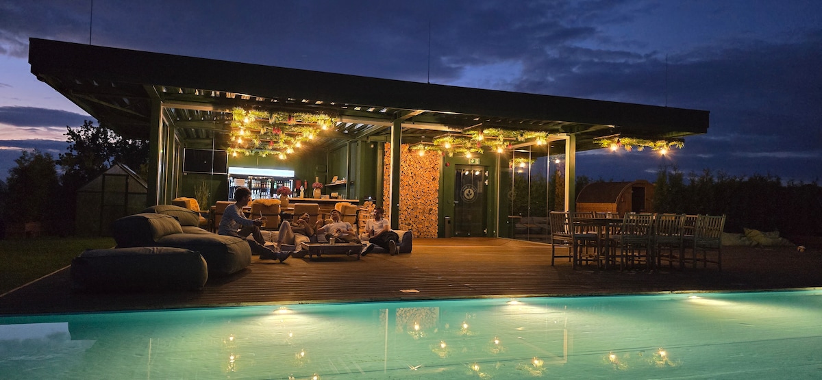An outdoor space features a bright pool lit with soft blue light, surrounded by seating areas. Twinkling string lights hang above a stylish patio, inviting relaxation as the evening sky transitions into twilight.