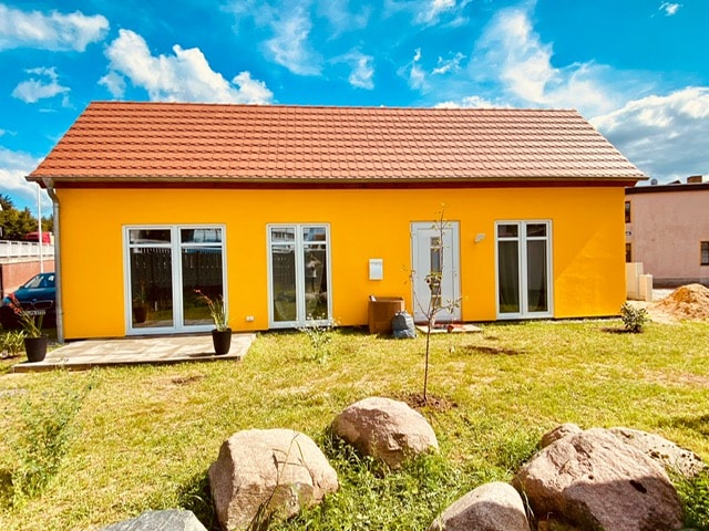 A bright yellow house is showcased against a blue sky, featuring large glass doors that provide natural light. A grassy area surrounds the property, with several rocks and potted plants positioned strategically outside. The exterior exudes a sense of openness and warmth.