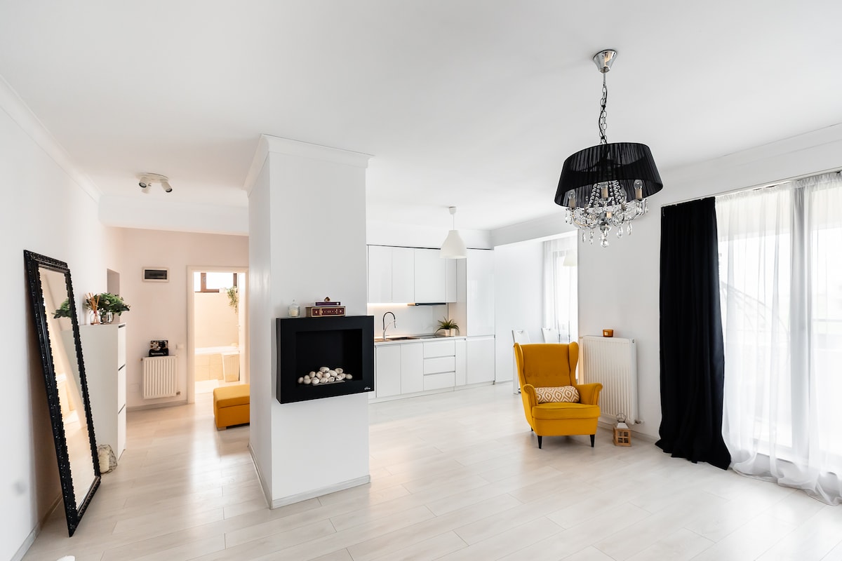 A spacious living area features a yellow armchair and a modern black chandelier above. A minimalist kitchen is visible in the background, with light cabinetry and natural light flowing through large windows. A decorative mirror and an electric fireplace add visual interest.