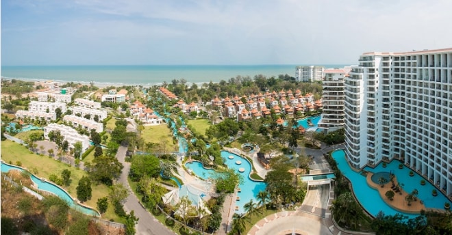 A panoramic view captures the resort, showcasing lush green landscapes interspersed with winding pools. The coastline is visible in the distance, framed by a clear blue sky. Multiple buildings are arranged harmoniously throughout the area, creating a serene atmosphere for relaxation.