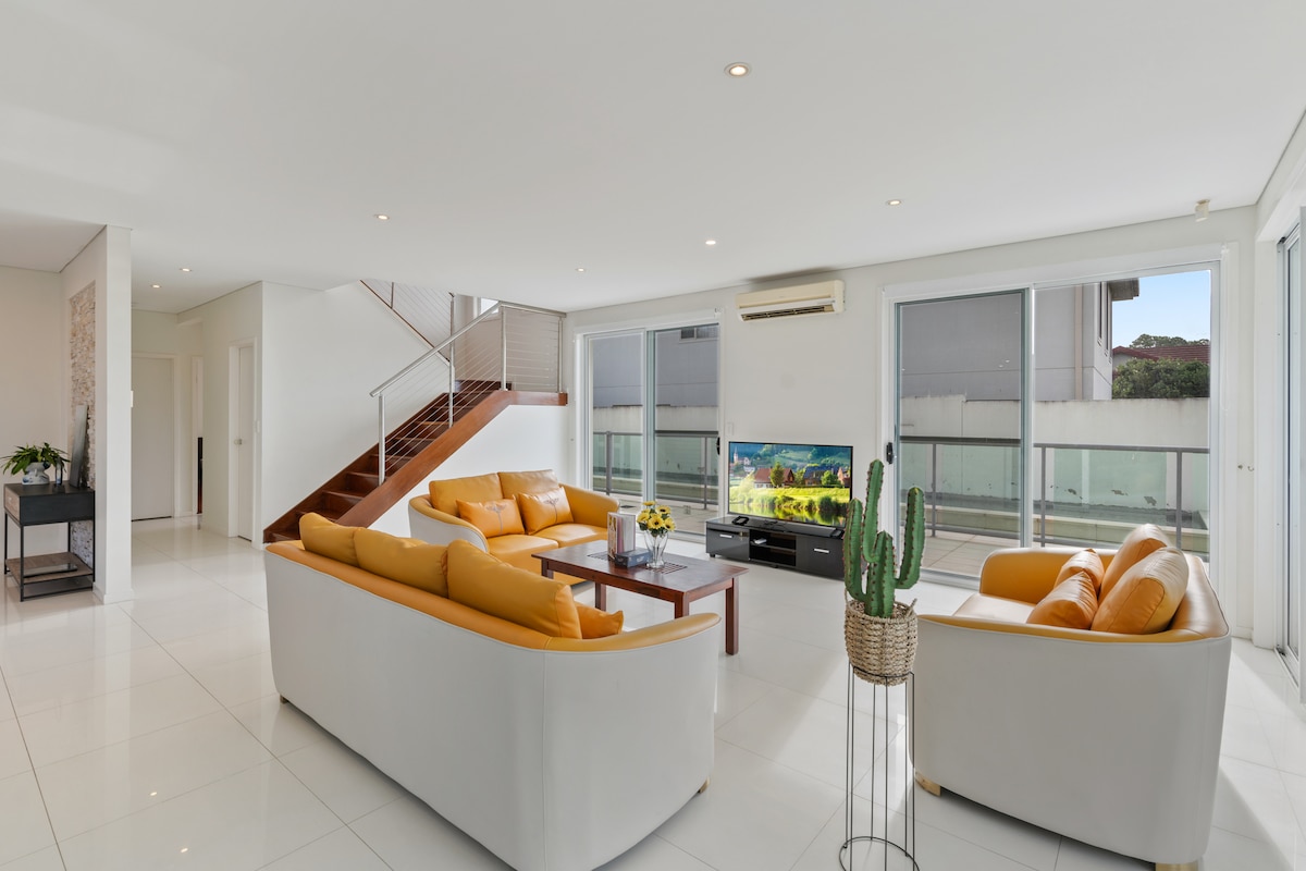 A spacious living area is showcased, featuring two comfortable seating arrangements in neutral colors. A television is positioned for viewing, with large glass doors allowing natural light and access to the outdoor space. A decorative cactus adds a touch of greenery.