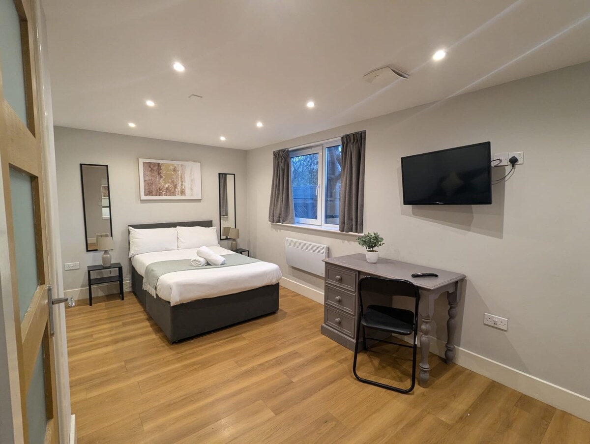 A well-lit studio apartment is showcased, featuring a spacious bed with crisp linens and a soft grey duvet. A desk is positioned near a window dressed with light curtains. Modern wall art and a flat-screen television complement the inviting decor.