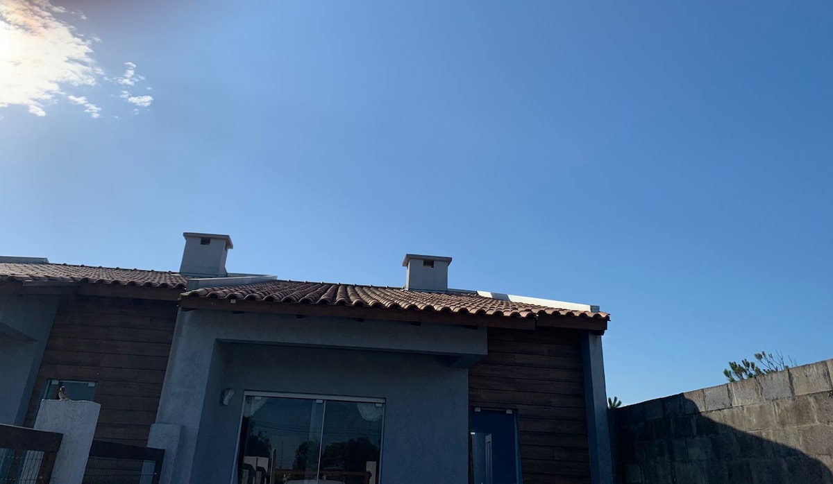 The exterior of the house is shown against a clear blue sky. The structure features a traditional tile roof and a combination of wooden and concrete finishes. Sunlight highlights the architecture, which includes chimney stacks and large windows.