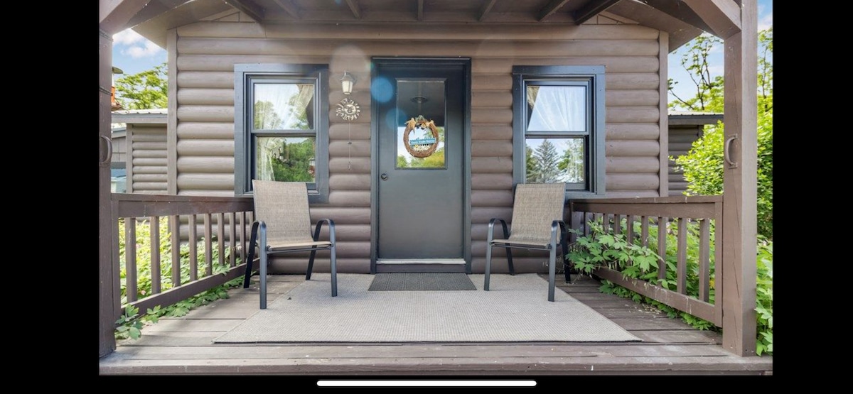 A welcoming front porch features two outdoor chairs positioned on a carpeted surface. A door adorned with a decorative wreath is framed by two windows, allowing natural light to enhance the entryway. Lush greenery surrounds the entrance.