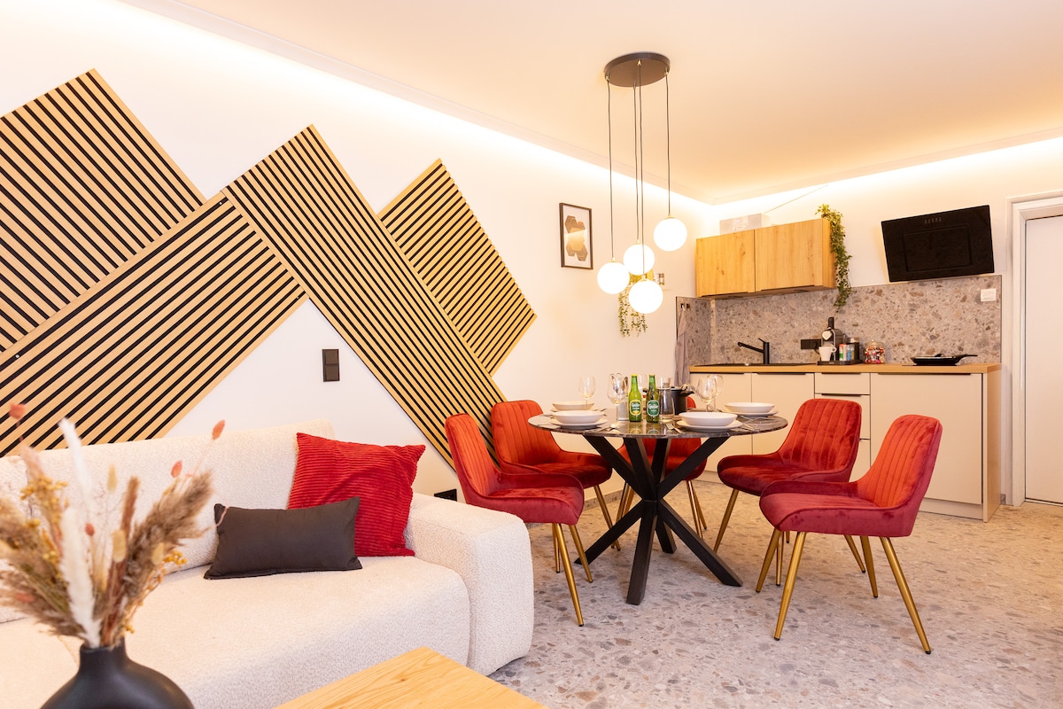 A modern living and dining area is highlighted by a round dining table with red chairs. A light-colored sofa is positioned in the foreground. The wall features dynamic acoustic panels, and warm lighting enhances the cozy atmosphere. A kitchenette is visible in the background.