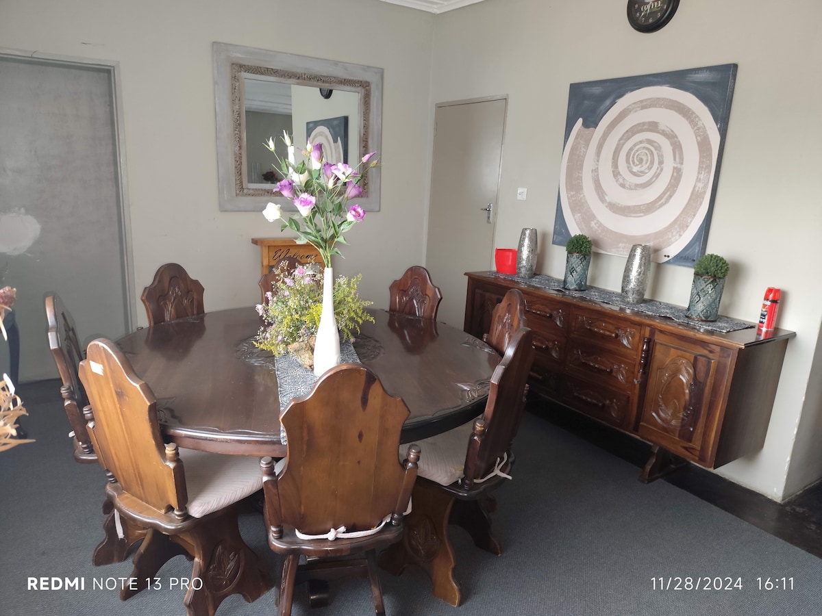 A wooden dining table with matching chairs is centered in the room, surrounded by a neutral-colored wall. A vase filled with flowers sits atop the table, complementing the wooden sideboard against the wall. A round mirror is positioned above the sideboard, adding depth to the space.