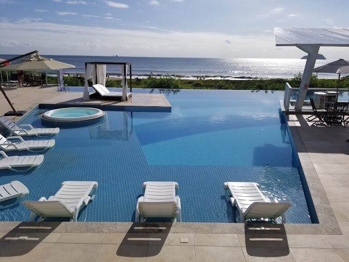 An expansive infinity pool is featured, reflecting the blue sky and ocean beyond. White lounge chairs are arranged around the pool area, alongside a raised hot tub. Umbrellas provide shaded seating spots, creating a tranquil outdoor environment.