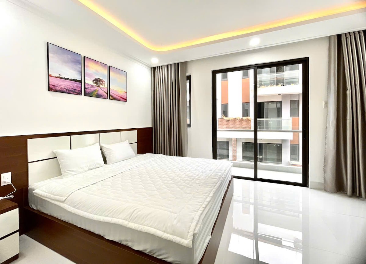 A bright sleeping area features a spacious bed with a white quilt. Large windows allow natural light to fill the room, while soft curtains frame the views of the outdoors. Three framed artworks with landscape themes are displayed on the wall.